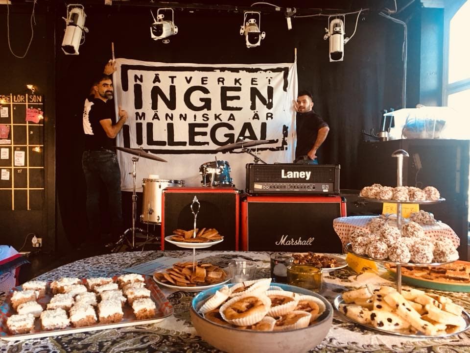 Two people setting up a 'No one is illegal' banner on a wall with fika in the front